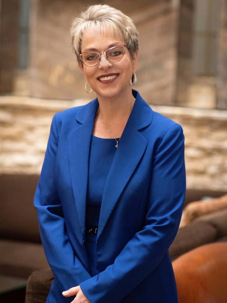 Image of Tina Brandau in a royal blue suit against a tan wall in a conference center